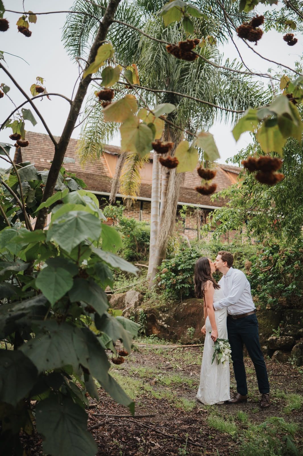 Letícia e Pablo se beijando no jardim, entre árvores e uma casa antiga